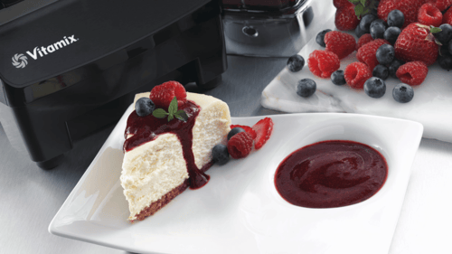 Close-up of bottom of Vitamix blender with logo. In front, white plate with cheesecake and sauce over it and berries behind.