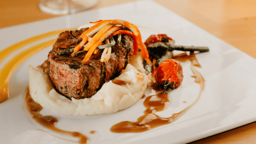 Small steak on top of mashed potatoes on white plate. Steak has thin slices of carrots and gravy on top.