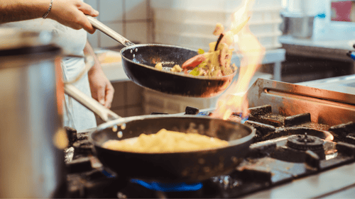 Two large resaurant-size fry pans are being held over two gas stove burners. Each fry pan has a long silver handle being held by a chef's hand. There is a large orange flame coming from underneath one pan. Each pan is full of food being tosses in the air.