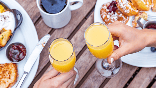 Two glasses of mimosas toasting with plates to the side and a cup of coffee at top of image